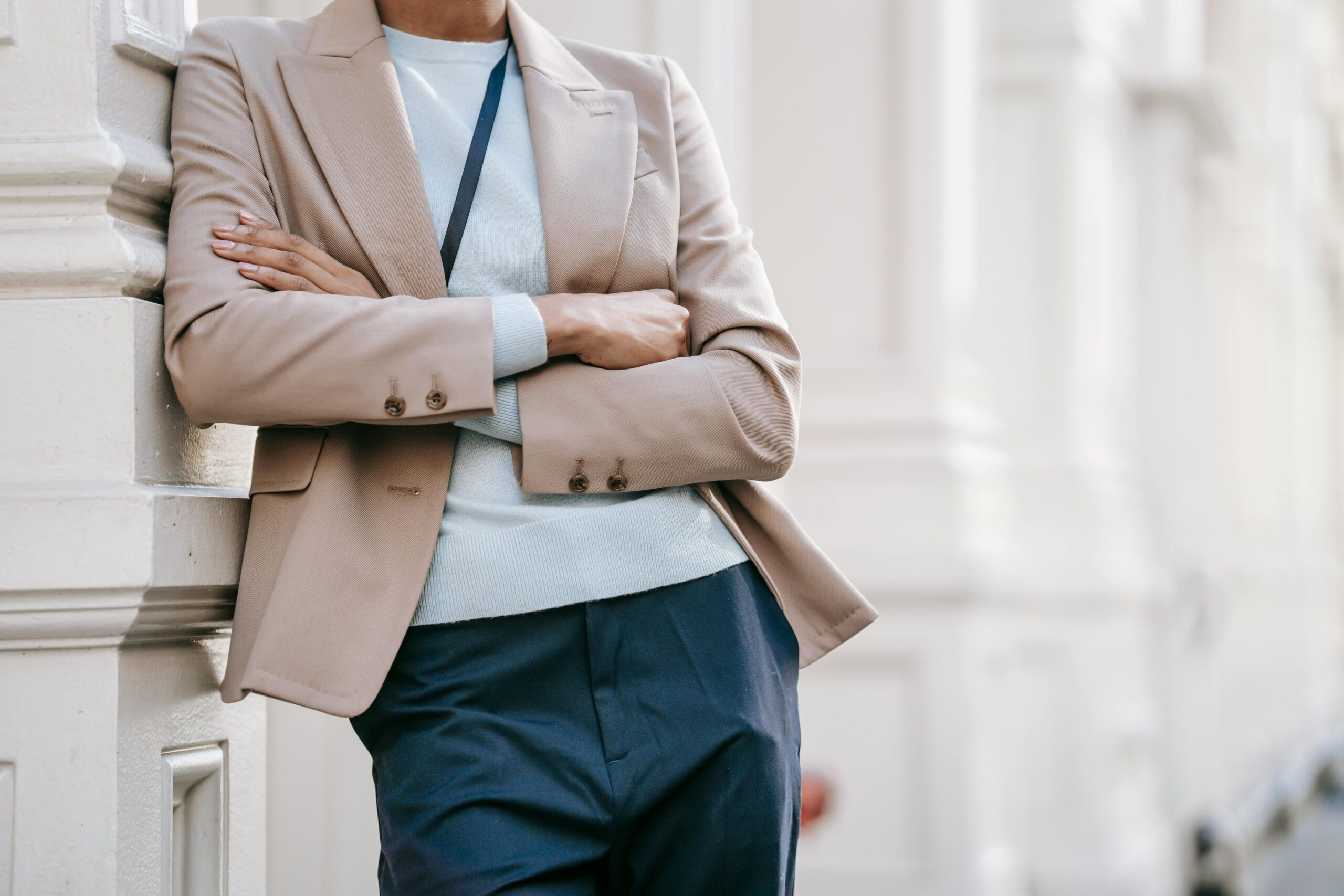 Stylish woman in a beige blazer and blue trousers leaning on a building wall with arms crossed.